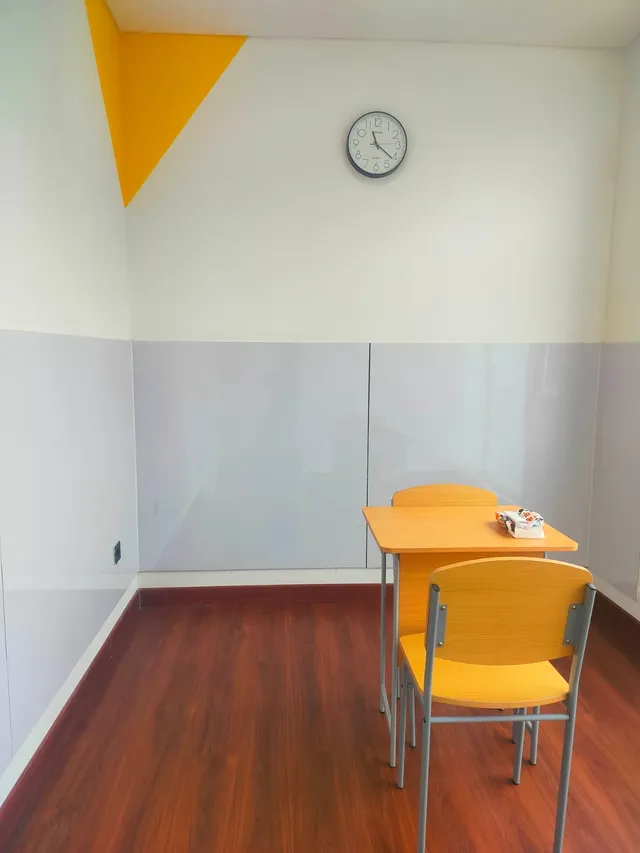 Room with a wooden table and two chairs, gray wall panels, and a wall clock